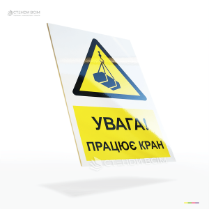 Табличка безпеки «Увага! Працює кран» поєднує універсальний знак загальної небезпеки з уточнюючим написом, що інформує про виконання робіт із використанням підйомної техніки.